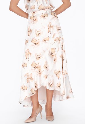 Zwiewna biała spódnica w beżowy kwiatowy wzór, z wysokim przodem i dłuższym tyłem, noszona z beżowymi szpilkami, ręce spoczywające na biodrach.