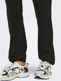 Personne portant un pantalon de jogging noir et des baskets blanches épaisses avec des détails noirs et argentés, debout sur un fond clair uni.