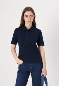 Marineblå polo med krage og korte ermer; har en knaplukking og en liten logo detalj på brystet. Mykt, glatt stoff.