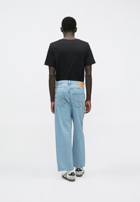 Hombre vistiendo una camiseta negra, pantalones vaqueros de pierna ancha de color azul claro y zapatillas blancas, de pie frente a un fondo blanco liso.