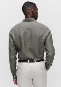 Olijfgroene linnen blouse met lange mouwen, rugpand en knoopdetails, gecombineerd met lichtgekleurde broeken en een gevlochten bruine riem.