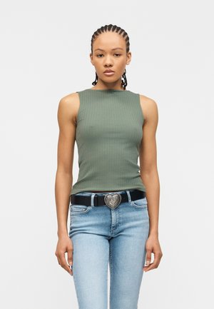 Γυναίκα με πλεξούδα, που φοράει αμάνικη πράσινη ριμπ πλεκτή μπλούζα, ανοιχτόχρωμα μπλε τζιν και μαύρη ζώνη με ασημένια αγκράφα σε σχήμα καρδιάς.