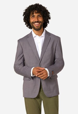 Mann mit lockigem Haar lächelt, trägt ein graues Sakko, weißes Hemd und olivgrüne Hose, steht mit verschränkten Händen.