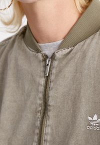 Olive green bomber jacket with ribbed collar and zipper closure. Features embroidered logo on left side, made from smooth fabric.