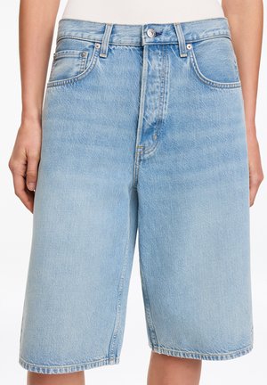 Hellblaue Jeansshorts mit vorderem Knopf und Reißverschluss, Fünf-Taschen-Design, getragen von einer stehenden Person mit entspannt an den Seiten hängenden Händen.