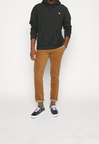 Felpa con cappuccio verde scuro con tasca, pantaloni in velluto a coste color sabbia e sneakers multicolori con lacci bianchi e dettagli di cucitura visibili.
