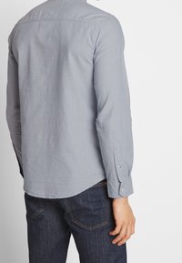 Vaaleanharmaa pitkähihainen paita, jossa on kaulus, napilliset hihansuut ja kaareva helma, yhdistetty tummansinisiin farkkuihin. Päällä näkyy sileä kangaspinta.