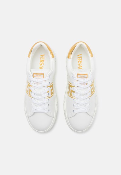 Fehér sneaker arany kiegészítőkkel, sima bőrből készült felsőrésszel, fűzős záródással és dombornyomott logó részlettel. Kerek orrú dizájn.
