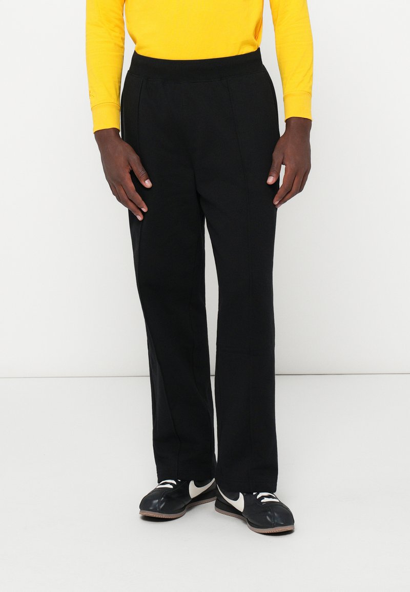 Persoană îmbrăcată cu pantaloni negri drepți, adidași negri și albi, și un tricou galben cu mânecă lungă, stând în fața unui fundal alb simplu.