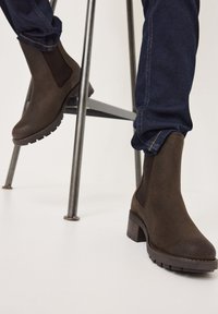 Stivali alla caviglia in suede marrone con pannelli laterali elastici, suola in gomma testurizzata e tacco a blocco, abbinati a jeans in denim scuro.