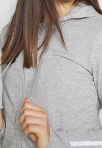 Felpa grigia con zipper e cordini. Realizzata in tessuto morbido, presenta una tasca e strisce a contrasto sulle maniche.