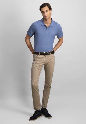 Mladý muž v modrém polotriku, béžových kalhotách, černém pleteném pásku a černých neformálních botách stojí před jednoduchým světle šedým pozadím.