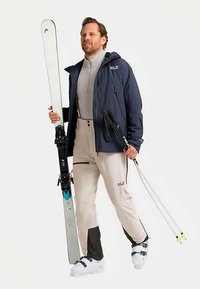 Skikleding bestaat uit een marineblauw jack, een grijze thermische top, beige broek met donkere accenten, witte skischoenen en witte ski's met zwarte bindingen.