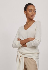 Helles Cremefarbiges V-Neck-Pullover aus weichem, gestricktem Stoff. Mit gerippten Bündchen und Saum. Getragen über hellbeigen Hosen mit einem gebundenen Detail.