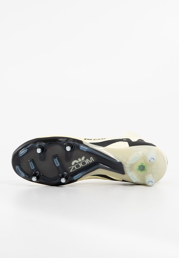 Chaussure à crampons avec une tige crème, des accents noirs et une semelle extérieure noire texturée comportant six crampons et un accent vert près du talon.