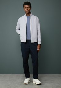 Man standing against gray wall wearing light gray zip-up jacket, blue cable-knit sweater, dark pants, and white sneakers with hands relaxed.