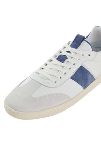 Sneaker bianco in pelle con dettagli in suede blu, punta rotonda, suola piatta beige e lacci. Presenta materiali lisci e testurizzati nel design.