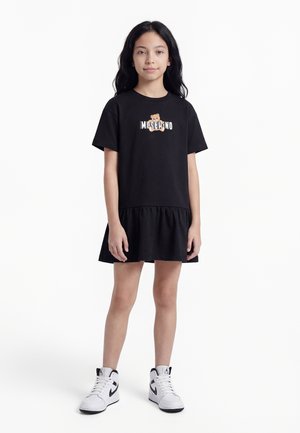 Κορίτσι που φοράει μαύρο φόρεμα με κοντά μανίκια, με λογότυπο αρκουδάκι και το κείμενο «Moschino», συνδυασμένο με λευκά και μαύρα ψηλά αθλητικά παπούτσια.