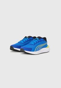 Une paire de chaussures de sport bleues avec des semelles noires, des lacets bleu clair et un détail de rayure jaune sur le côté, légèrement inclinée vers la droite sur un fond blanc.