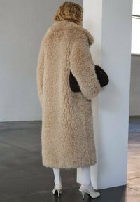 Cappotto in pelliccia sintetica beige con ampio colletto, maniche lunghe e silhouette dritta. La modella tiene una pochette marrone scuro, indossando collant bianchi e tacchi.