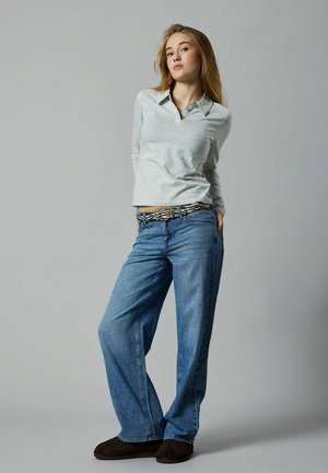 Ung kvinna som står med händerna bakom ryggen, iklädd en ljusgrå långärmad polo, blå jeans, mönstrat skärp och mörka tofflor, mot en grå bakgrund.