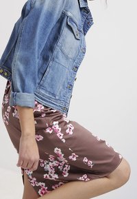 Denimjakke med lys vask, store frontlommer og brune knapper, båret over en brun kjole med blomsterprint i pink og hvid.