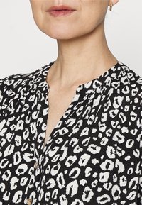 Schwarze Bluse mit weißem Leopardenmuster, ausgestattet mit einer Knopfleiste, gefälteltem Ausschnitt und einem weichen, leichten Stoff. 
