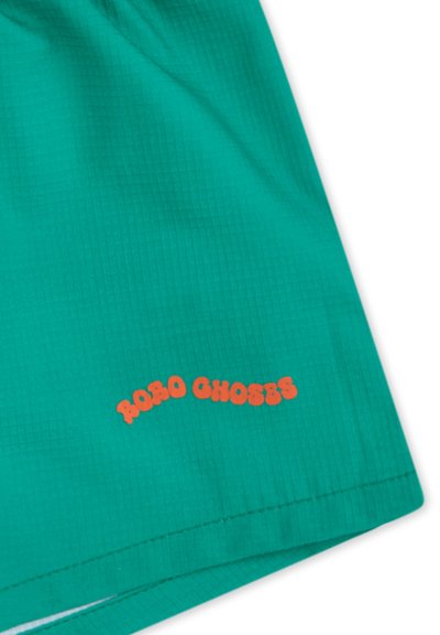Pantaloni corti verdi con una superficie testurizzata, dotati di un logo arancione "ROBO CHOSES" vicino all'orlo. Design semplice, tessuto leggero, stile informale.