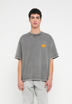 Hallikas ühevärviline ülimõõdus t-särk ümarate kaelakaare, lühikeste varrukatega ja tekstuurse kollase plaastriga rinnal. Stiilitud heledate hallide teksapükstega.