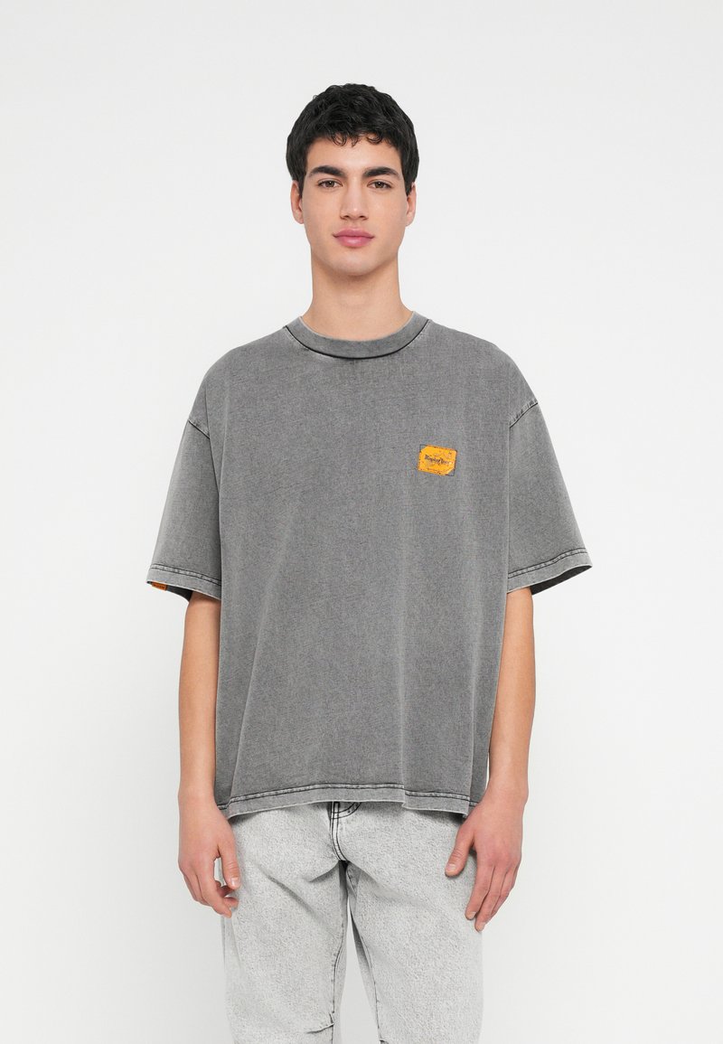 Γκρι φαρδύ t-shirt με στρογγυλή λαιμόκοψη, κοντά μανίκια και υφασμάτινη κίτρινη επιφάνεια στο στήθος. Συνδυασμένο με ανοιχτό γκρι τζιν παντελόνια.