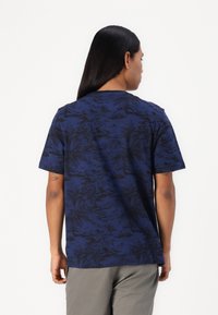 Tricou bleumarin cu un model tropical negru, mâneci scurte, decolteu rotund și croială lejeră. Materialul pare moale și ușor.