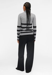 Pull gris tricoté avec des rayures horizontales noires, col montant et bord côte, associé à un pantalon noir ample. Matière en laine texturée.
