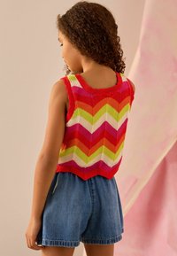 Gebreide mouwloze top met een gegolfde halslijn, versierd met een zigzagpatroon in rood, roze, oranje, geel, groen en wit; gecombineerd met denim shorts.
