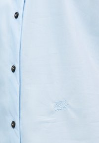 Ανοιχτό γαλάζιο πουκάμισο από ύφασμα με μαύρα κουμπιά και ένα διακριτικό κεντητό λογότυπο σε λευκό στο στήθος. Λεία υφή και κλασικός σχεδιασμός.