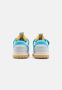 Sneaker con una parte superiore grigio chiaro, accenti blu e fodera gialla; presenta una suola beige spessa e un manico sul retro per facilitare l'indossamento.