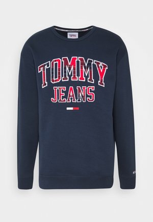 Granatowa bluza z długim rękawem z "TOMMY JEANS" dużymi czerwonymi i białymi literami oraz małym czerwono-białym logo flagi poniżej.