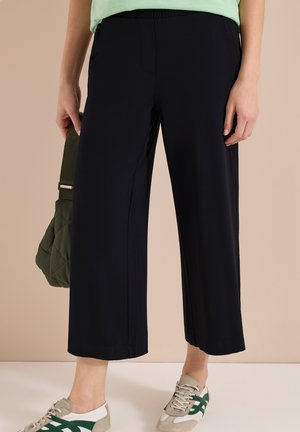 Persona vistiendo pantalones negros anchos y cortos, sosteniendo un bolso de mano verde acolchado, y zapatillas beige con rayas verdes sobre un fondo liso.