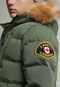 Zelena puffer jakna koja ima uklonjivu kapu obloženu krznom, crno-zlatni iverak s oznakom "Zavetti Canada" i prošivenu teksturu.