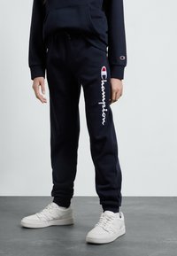 Pantalons de survêtement navy en mélange de coton, avec un logo "Champion" brodé en blanc et un logo d'accent sur la hanche, associés à des baskets blanches.