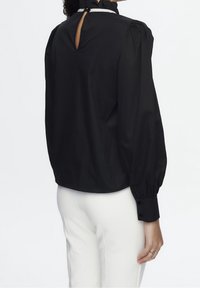 Zwarte blouse met opbollende mouwen, hoge halslijn en sleutelgatdetail aan de achterkant, gemaakt van een gladde stof, gecombineerd met witte broeken.