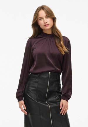 Blusa di satin bordeaux con maniche lunghe e scollo arricciato, abbinata a una gonna in pelle nera a vita alta con chiusura zip frontale.