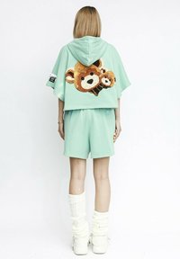 Sudadera corta de color verde menta con un gran diseño de oso de lentejuelas en la parte posterior, combinada con pantalones cortos a juego y calentadores de piernas de canalé blancos.