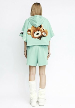 Sudadera corta de color verde menta con un gran diseño de oso de lentejuelas en la parte posterior, combinada con pantalones cortos a juego y calentadores de piernas de canalé blancos.