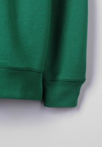 Grön bomullströja i tyg med ribbade ärmslut, som har en slät textur och en enfärgad yta, med en enkel design och utan ytterligare mönster.