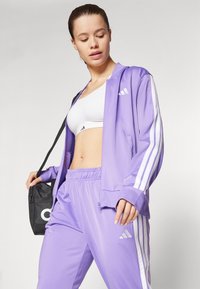 Šviesiai violetinė sportinė apranga su užtrauktuku, balto juostelėmis ir atitinkamomis kelnėmis. Po ja matosi balta sportinė liemenėlė.