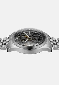 Srebrny metalowy zegarek na rękę z szkieletowym cyferblatem, rzymskimi cyframi, widocznymi zębatkami i bransoletą na ogniwa na białym tle.