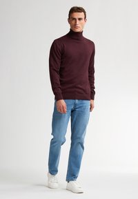 Burgunderfarbener Rollkragenpullover mit langen Ärmeln, kombiniert mit hellblauen Jeans und weißen Sneakers, vor einem schlichten Hintergrund stehend.