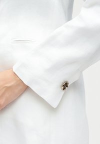 Λευκό μανίκι σακακιού με ασημένια λεπτομέρεια κουμπιού στο μανσέτα, που ακουμπά κοντά σε πλαϊνή τσέπη, φορεμένο στο χέρι ενός ατόμου.