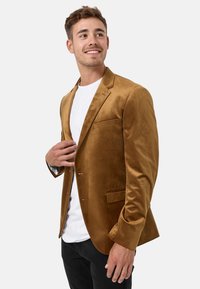 Blazer de terciopelo marrón con cierre de un solo botón, solapas de muesca y un bolsillo en el pecho, llevado sobre una camiseta blanca y pantalones negros.