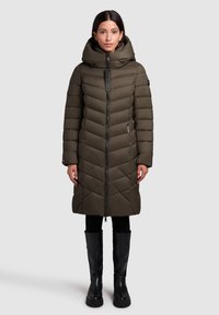 Olīvzaļš izšūts mētelis ar chevron šūšanas dizainu, augstu apkakli un ar rāvējslēdzēju; apvienots ar melniem gumijas zābakiem līdz ceļgaliem.